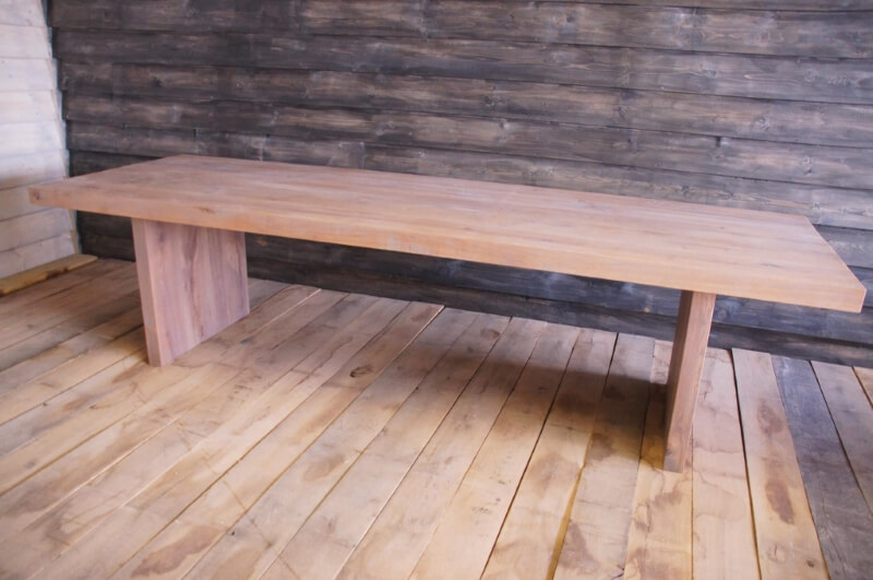 Lange, rechthoekige eikenhouten tafel met een lichte, natuurlijke afwerking en brede poten, geplaatst in een ruimte met een houten vloer en donkere houten achtergrond.