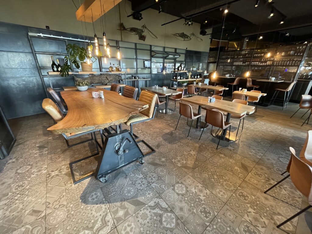 Organisch gevormde walnoothouten bartafel met industrieel zwart metalen onderstel, omringd door leren barkrukken in een sfeervolle ruimte met decoratieve tegelvloer.