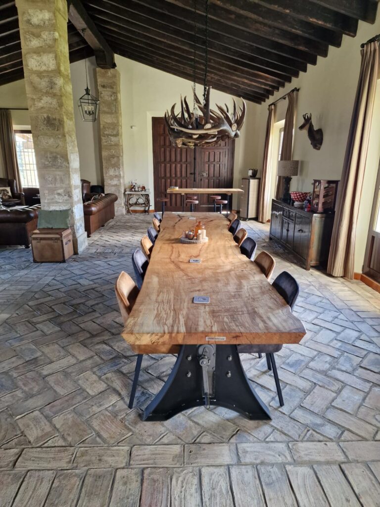 Industriële eettafel met een massief houten blad en een robuust zwart stalen onderstel, omringd door moderne stoelen op een betegelde vloer in een ruime eetkamer.
