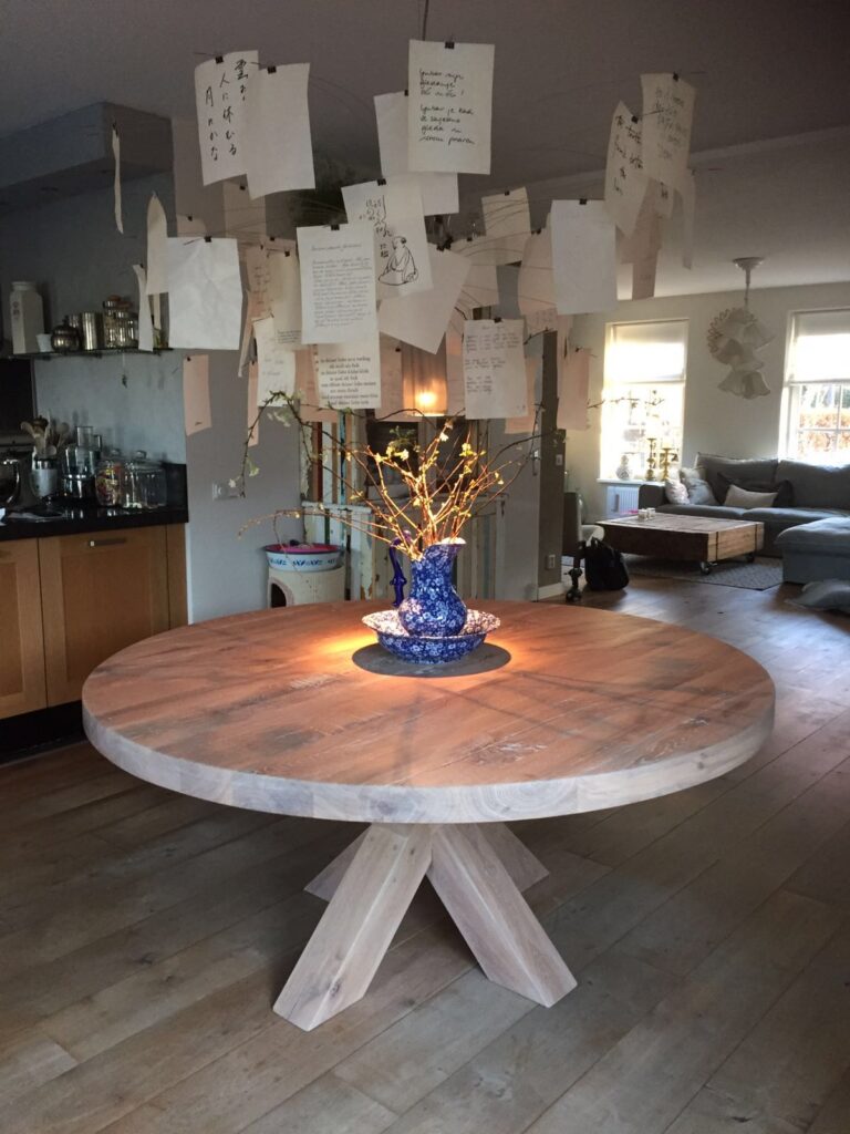 Ronde eikenhouten tafel met een matte afwerking en kruislings geplaatste poten, gedecoreerd met een blauwe vaas en takken in een huiselijke woonruimte.